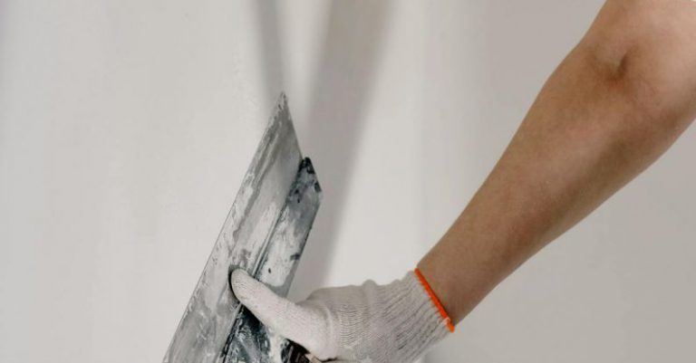 Contractor Vs DIY - Crop anonymous male worker in gloves holding putty knife while working at home