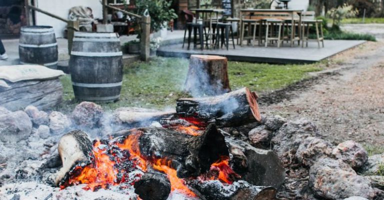 Backyard Fire Pit - Stone fire pit with burning firewood placed on courtyard of residential building with chairs and table on terrace in countryside