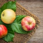 Three ripe apples on leaves over a rustic wooden background, emphasizing freshness and nutrition.